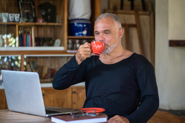 Mature man freelancer working with laptop and phone holding red cup of coffee