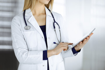 Unknown woman-doctor is holding a tablet computer in her hands, while standing in a clinic cabinet. Female physician at work, close-up. Perfect medical service in a hospital. Medicine concept