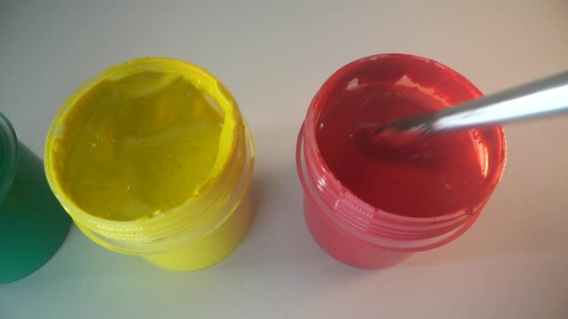 Green, redd and yellow paint cans on the white surface. Painter dips the blush in red color. Top view. Macro shot
