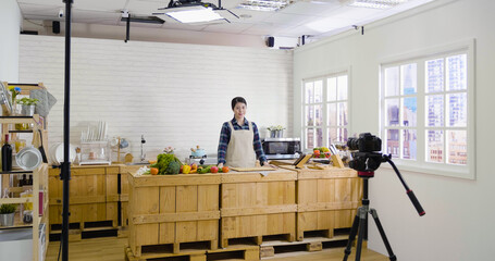 Fototapeta na wymiar beautiful young asian woman filming video blog episode about healthy food cooking while standing at kitchen in studio. female presenter on cookery show talking to professional camera with tripod