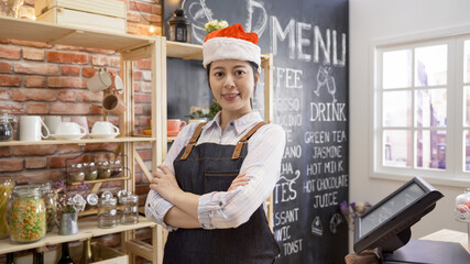 Portrait of confident young woman worker wearing apron and red santa hat standing behind cafe counter. happy female barista looking at camera and smiling enjoy xmas. waitress work in christmas