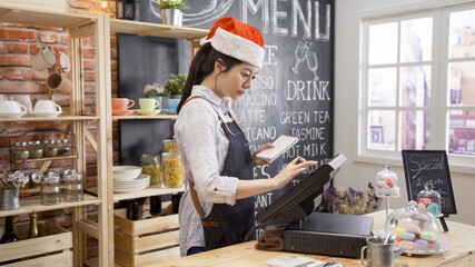 waitress in santa hat and apron prepares bill on computer POS tablet pc in counter bar of cafeteria. young girl barista working in christmas coffee shop. cafe staff touching screen on sale terminal.