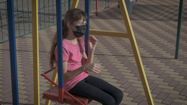 Quarantined girl on swing. A sad teen in medical mask sits and think in the swing.
