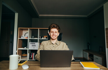 Happy guy in wireless headphones works at home on the Internet at his desk, looks at the camera and smiles. Portrait of a joyful programmer working remotely at the computer in a cozy room at home.