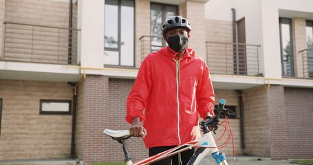 Portrait of African American young deliveryman in casque standing at his bicycle at street.