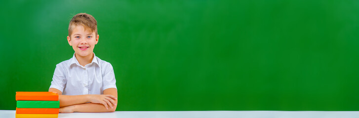 The boy sits on the background of a school blackboard next to textbooks. Horizontally stretched panoramic image for banner