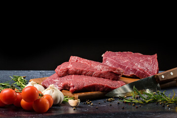 Three pieces of beef steak with a knife, rosemary, tomatoes, spices and garlic on a black background with copy space.