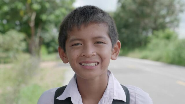 The slow motion of smiling poor elementary school boy in Thai elementary school uniform wait a bus to go back home after school evening beside the road in the countryside of Thailand. 