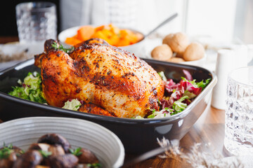 Whole roasted chicken served with fresh salad in black pan on a festive table. Thanksgiving or family dinner celebration cooking concept.