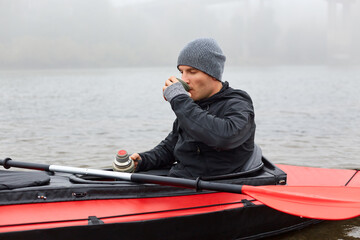 Breakfast in canoe in middle of river, man enjoying hot beverage from thermos, guy wearing warm black jacket and gray cap drinks coffee or tea in kayak.