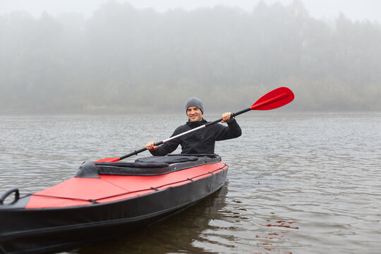 Man sitting smiling in kayak and holding paddle in hands, sportsman wearing black jacket rowing boat in foggy morning, water sport in cold autumn.