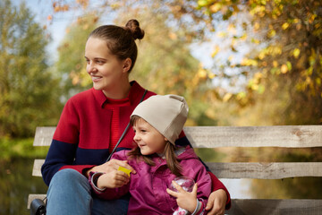 Young dark haired mother with daughter sitting on wooden bench in autumn park near lake or river,...