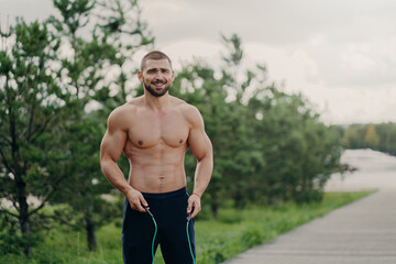 Cheerful unshaven sportsman skips with rope, has muscular body, enjoys fitness training, poses outdoor. Strong man with naked torso uses sport equipment for workout, breathes fresh air in nature