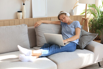 Smiling young woman with headphones and laptop on the sofa.