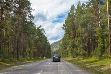 Asphalt road in Altai mountains on summer day against background of hilltops and green coniferous forest with an SUV car moving through the trees. Free path during a vacation outside city in nature.