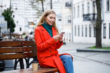 Cheerful young woman tapping and scrolling on mobile phone and sitting on bench outside. Stylish attractive female messaging on smartphone. Telephone chatting.