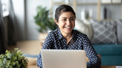 Happy cheerful millennial indian business woman sitting at desk in front of laptop laughing talking...
