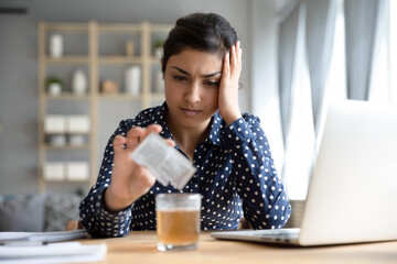 Indian woman sit at office desk touch head pouring into glass of water anti hangover powder revival remedy. Abuse excessive alcohol consumption, seasonal grippe virus treatment need sick leave concept