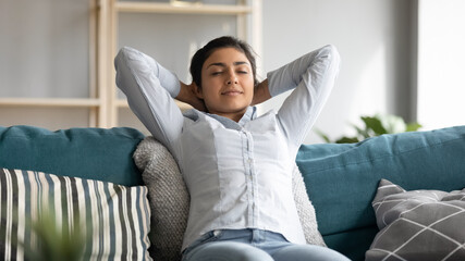 Serene Indian woman closed eyes breath fresh humified air in cozy living room alone put hands...