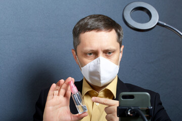 A man in a suit without a tie. Medical mask on the face. Demonstrates a spray bottle with an antiseptic on the camera. In front of him is a stand for a smartphone with a lamp. Video communication.