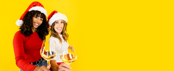 Fototapeta premium Toasting to Christmas together regardless of diversity - Two women, black Hispanic and Caucasian wearing Santa hat, looking at the camera holding a glass of white wine on big yellow copy space