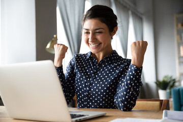 Overjoyed indian ethnicity girl sit at desk looks at laptop screen read incredible news clench...