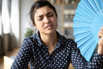 Overheated indian woman sit at workplace without air conditioner hold blue handheld fan cools...