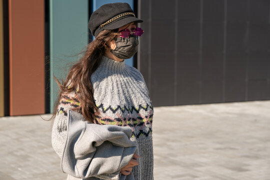 A Girl In A Cap And Pink Glasses Stands On The Street With A Black Protective Mask On Her Face. See Through Rose-tinted Glasses