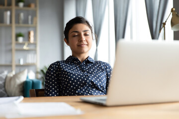 Serene indian business woman smiling sit at desk lean on office chair, enjoy break pause after working hard closing eyes daydreaming relaxing, feels satisfaction after project accomplishment concept
