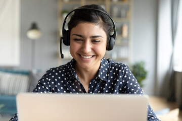 Cheerful Indian woman e tutor sit at desk home office wear headset looks at laptop talk by video call, helpline employee and pleasant chat to client remotely, distance webinar online teaching concept