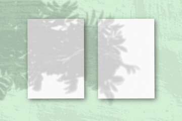 2 vertical sheets of textured white paper on soft greentable background. Mockup overlay with the plant shadows. Natural light casts shadows from an exotic plant. Horizontal orientation