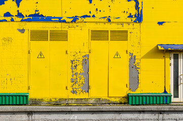 Doors with high voltage signs leading to technical rooms on the facade of a building with peeling yellow paint and visible spots of blue paint.