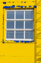 A window in a white frame on the front of the building, painted with peeling yellow paint and patches of blue paint.