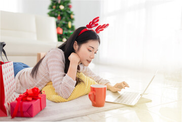 attractive asian female shopping with online market in Christmas celebration seasonal, she rest face on hand and stay on floor, she search and play internet with laptop, she feeling happy and smile