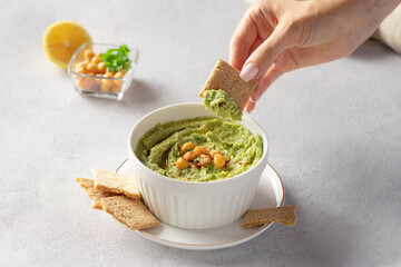 Female hand dipping cracker into white bowl with avocado hummus. healthy vegan dip. close-up. gray...