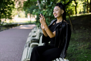 Obraz na płótnie Canvas Lovely cheerful young Asian brunette woman, wearing trendy black outfit, sitting on the bench at the city park, holding mobile phone, while reading funny memes or message