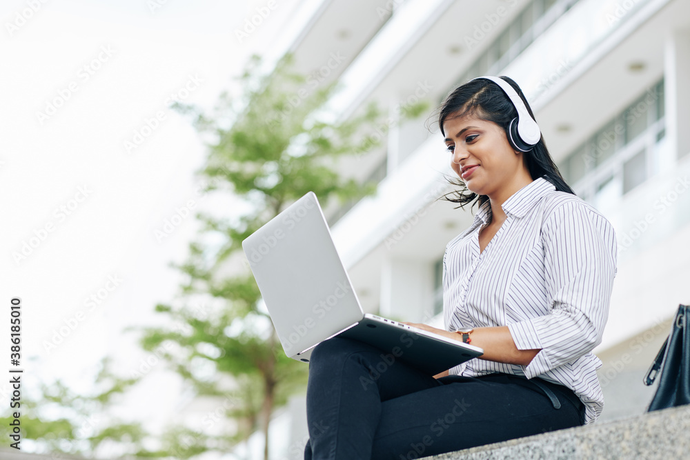 Wall mural smiling pretty indian businesswoman in headphones working on laptop outdoors