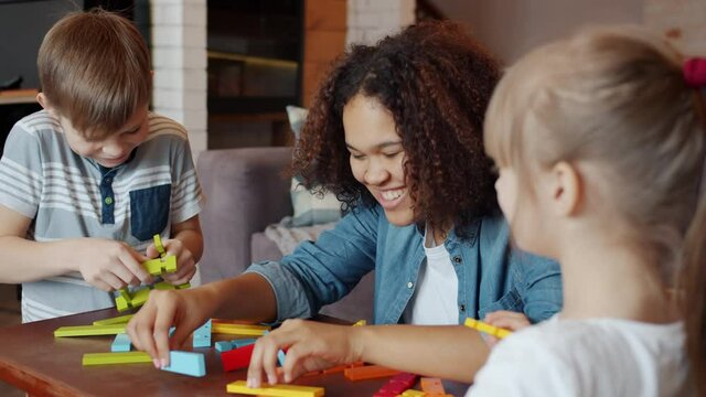 African-American girl nanny is talking to happy children and playing with bright wooden construction blocks in apartment. Games and childcare concept.