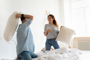 Happy laughing young couple playing pillow fight in bedroom, smiling beautiful woman and man having...