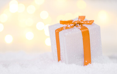 christmas gift box with snowflakes.

Christmas gift box in silver color with orange ribbon and snowflakes on the right against a background of blurry lights with space for text on the left, close-up s