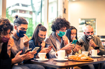 Multiracial friends with face mask holding smartphones sitting at table drinking cappuccino at cafe shop in the covid-19 time