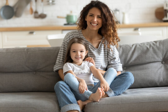 Portrait smiling young mother hugging little daughter, looking at camera, positive happy beautiful woman mum and cute adorable girl kid sitting on couch at home, posing for photo together