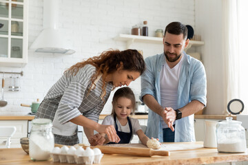 Happy parents and adorable little daughter cooking dough together, standing in modern kitchen, young mother and father teaching little girl to bake biscuits or muffins, family enjoying leisure time
