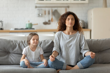Peaceful young mother and little daughter meditating, doing yoga exercise, sitting in lotus pose...