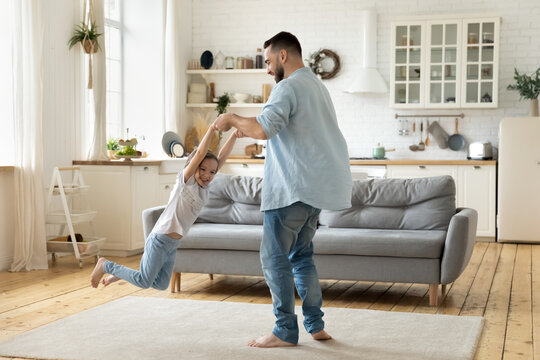 Happy young father spinning little daughter, holding hands, family playing funny active game at home, overjoyed dad and laughing cute preschool girl having fun in modern living room together