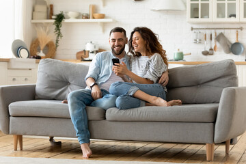 Happy young woman and man hugging, using smartphone together, sitting on cozy couch at home, smiling overjoyed wife and husband looking at phone screen, sitting on sofa in modern living room - obrazy, fototapety, plakaty