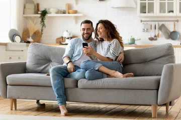 Fotobehang Happy young woman and man hugging, using smartphone together, sitting on cozy couch at home, smiling overjoyed wife and husband looking at phone screen, sitting on sofa in modern living room © fizkes