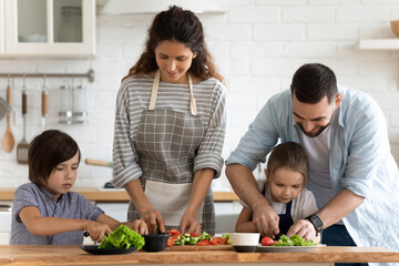 Happy young family with two adorable little kids cooking salad together, caring father teaching...