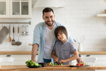 Family portrait smiling father and little son cooking dinner together, standing at wooden countertop in modern kitchen, happy dad with adorable kid preparing vegetables for salad, enjoying weekend