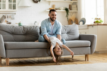 Loving young father playing with daughter at home, happy dad holding cute little girl upside down, sitting on cozy couch in living room, family playing funny game, spending leisure time together
